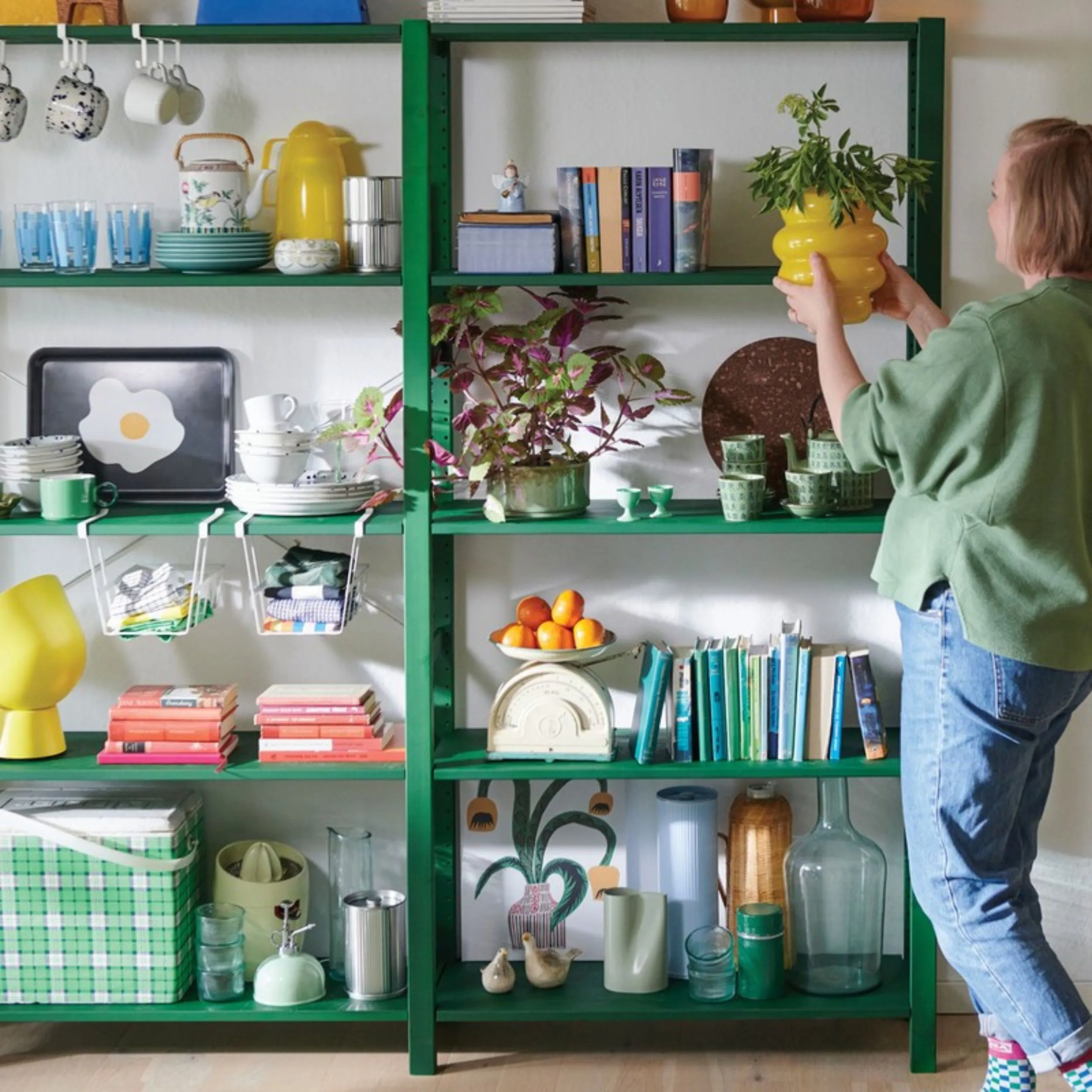 Étagère verte IKEA avec vaisselle et livres, une femme plaçant un pot jaune